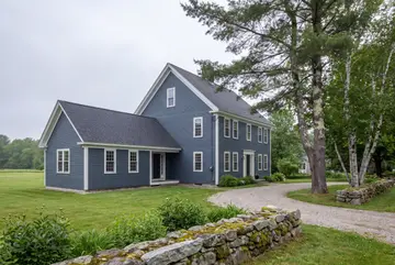 home addition builder: illustrative scene, A blue farmhouse with a new addition, seen from a three-quarter exterior view, surrounded by green lawn, trees, and a stone wall