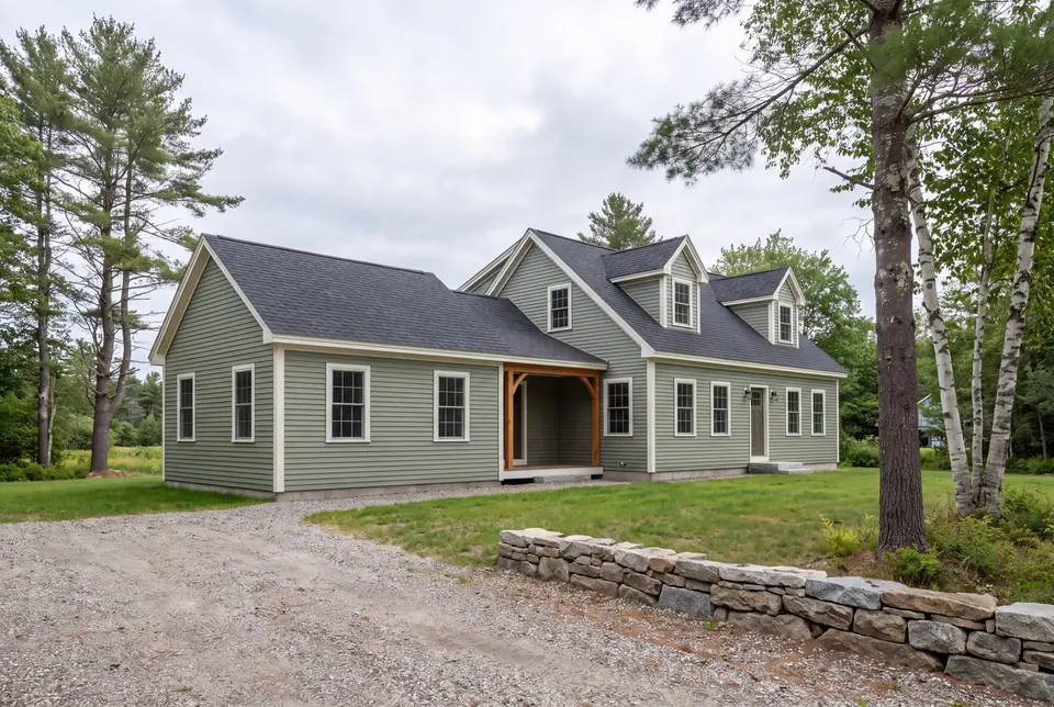 Illustration: A sage green house with a dark roof and white trim, featuring a main section with dormers and a single-story addition, seen from a three-quarter exterior view with a gravel driveway and trees