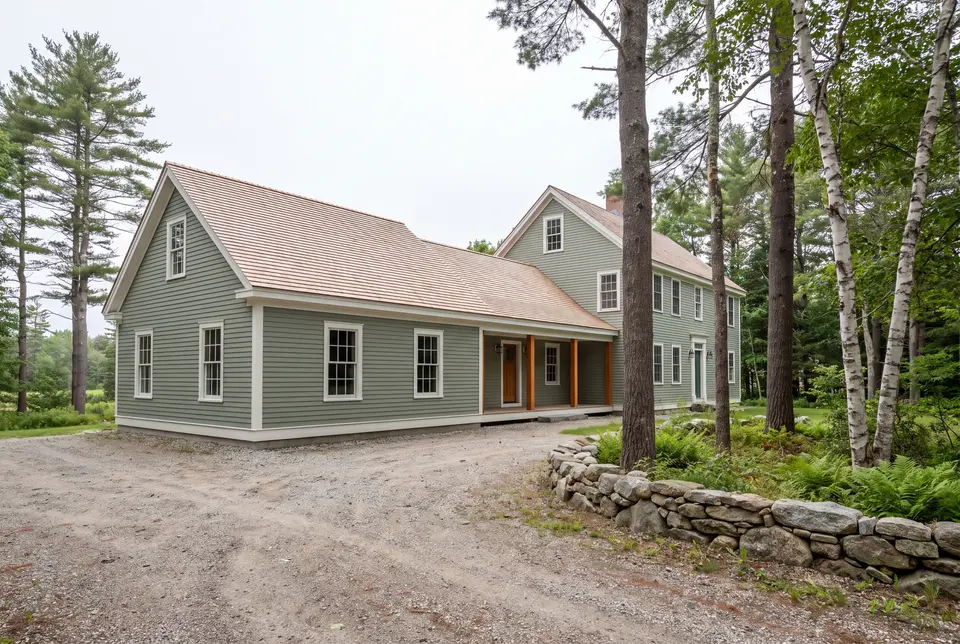 Illustration: A sage green clapboard house with a new single-story addition, light brown shingle roofs, and white trim, viewed from a three-quarter exterior perspective with a gravel driveway and surrounding trees