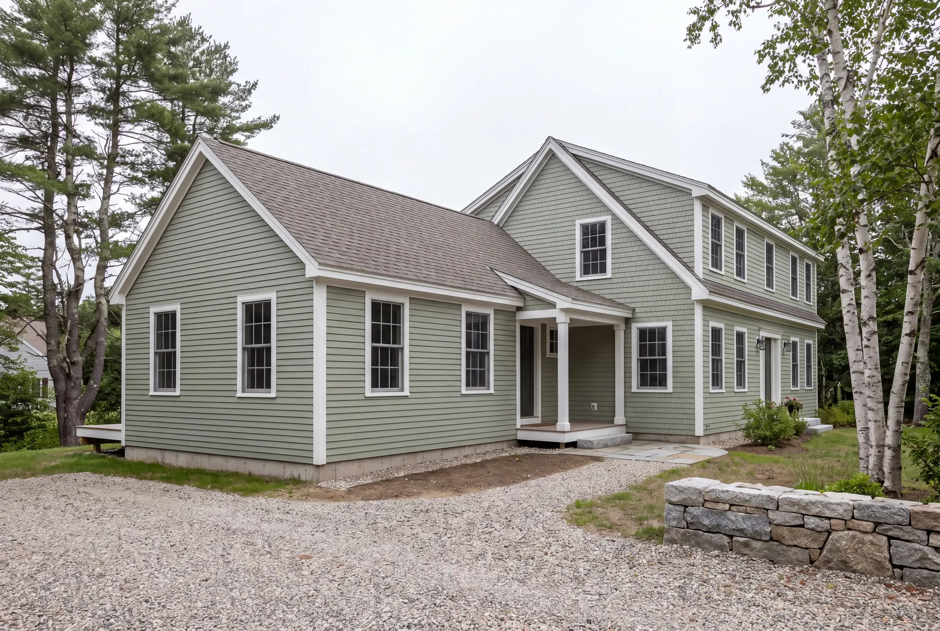 Illustration: A sage green clapboard house with a new single-story addition, viewed from a three-quarter exterior angle, showing matching architectural details and a gravel driveway