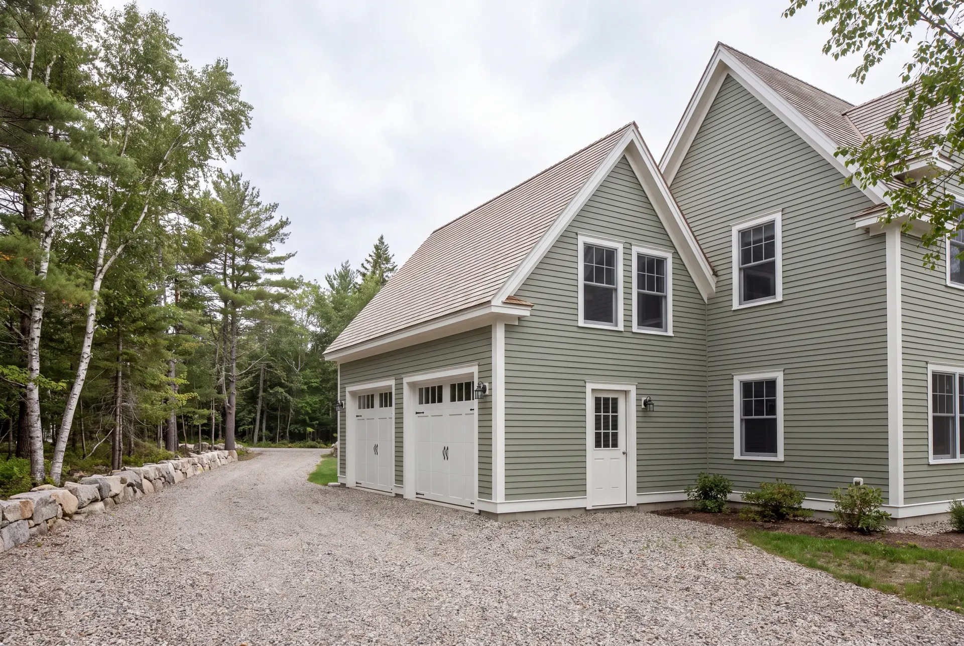 Illustration: A newly built sage green house with an attached two-car garage, featuring white doors and trim, a light brown roof, and a gravel driveway