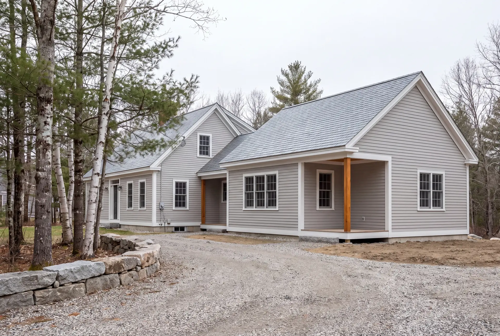 Illustration: A light gray clapboard house with a new addition, featuring a covered entry with wooden posts, a gravel driveway, and surrounding trees under a gray sky