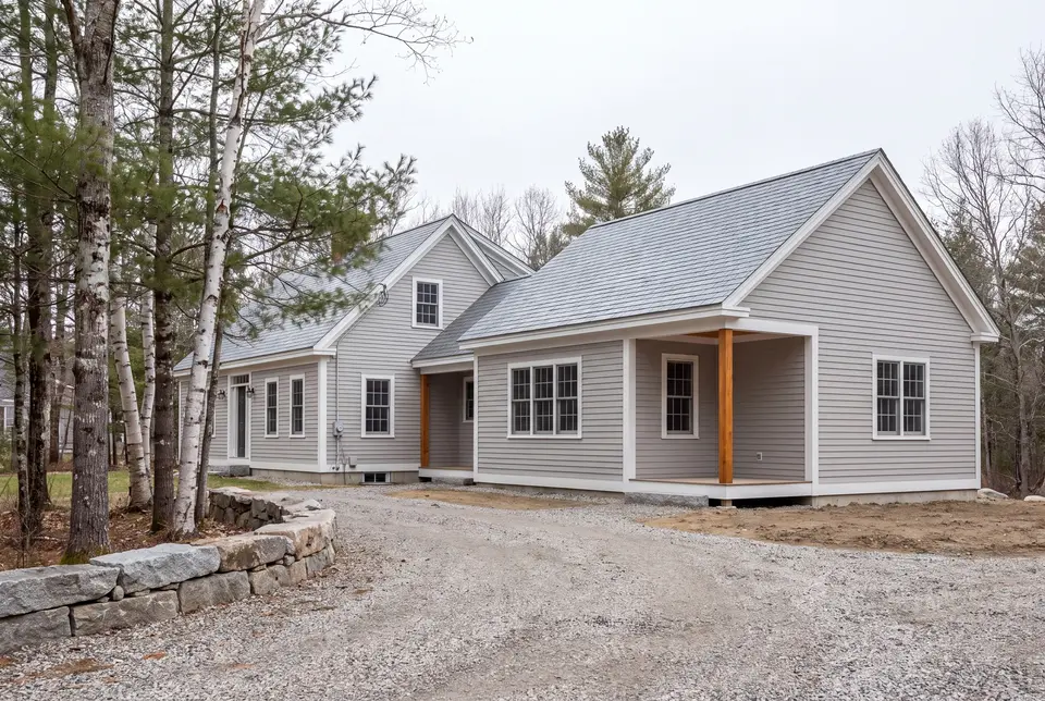 Illustration: A light gray clapboard house with a new addition, featuring a covered entry with wooden posts, a gravel driveway, and surrounding trees under a gray sky