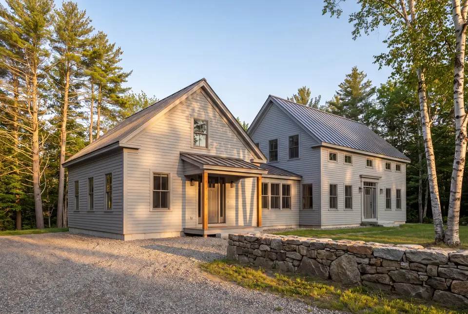 Illustration: A newly constructed single-story gray clapboard addition connected to an existing two-story gray clapboard house, both with dark gray metal roofs, seen from a three-quarter exterior view with a gravel driveway and stone wall in the foreground
