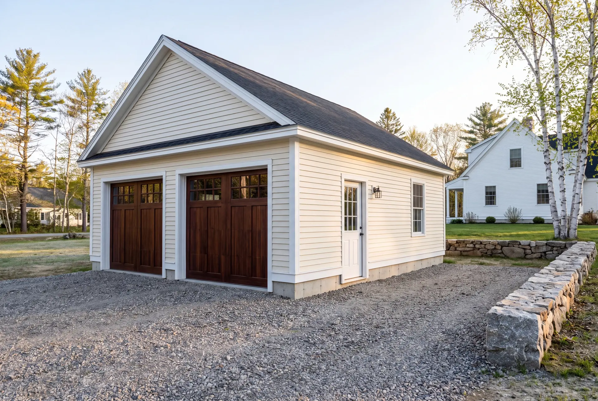 Illustration: A newly built, detached two-bay garage with cream siding and dark brown doors, situated next to a white house with a gravel driveway and a stone wall