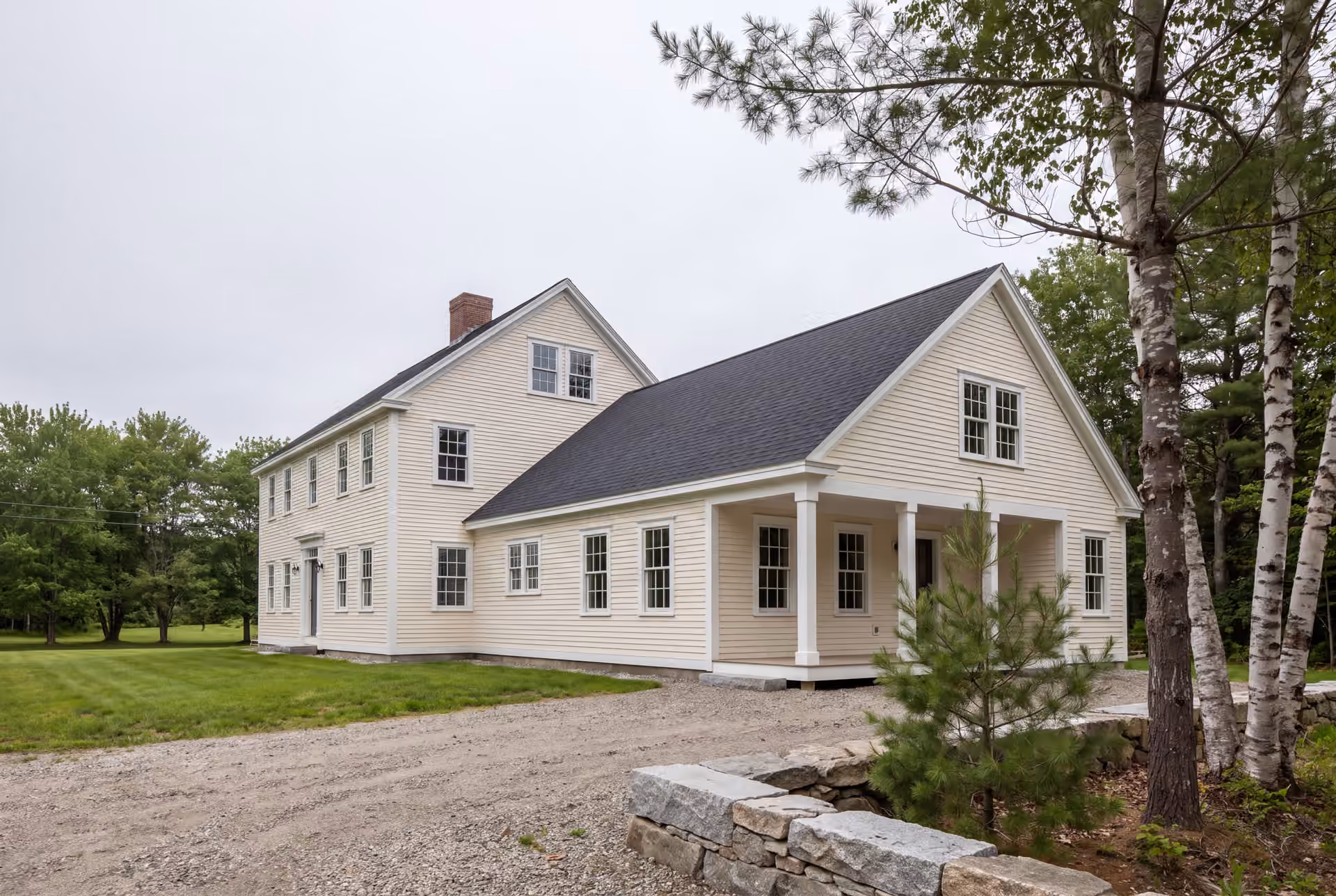 home addition builder in Cumberland, ME: illustrative scene, A cream-colored Colonial house with a new single-story addition, featuring a covered porch, seen from a three-quarter exterior view with a gravel driveway and trees