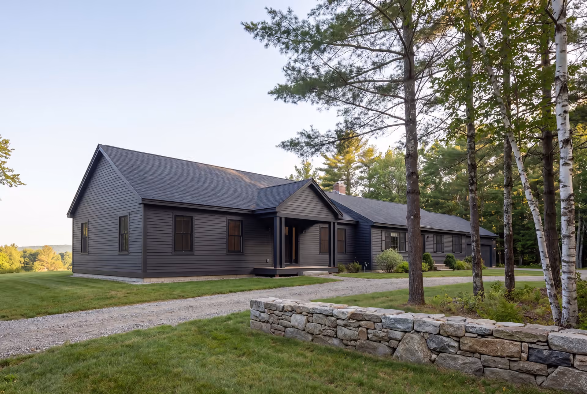 home addition builder in Dayton, ME: illustrative scene, A dark charcoal single-story house with a gabled entry porch, a gravel driveway, and a stone wall in the foreground, surrounded by trees and a grassy landscape