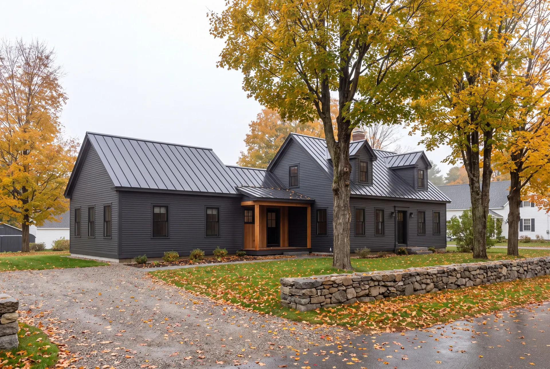 Illustration: An exterior three-quarter view of a dark charcoal house with a new single-story addition, featuring a metal roof and a wooden covered entry, surrounded by autumn trees and a stone wall