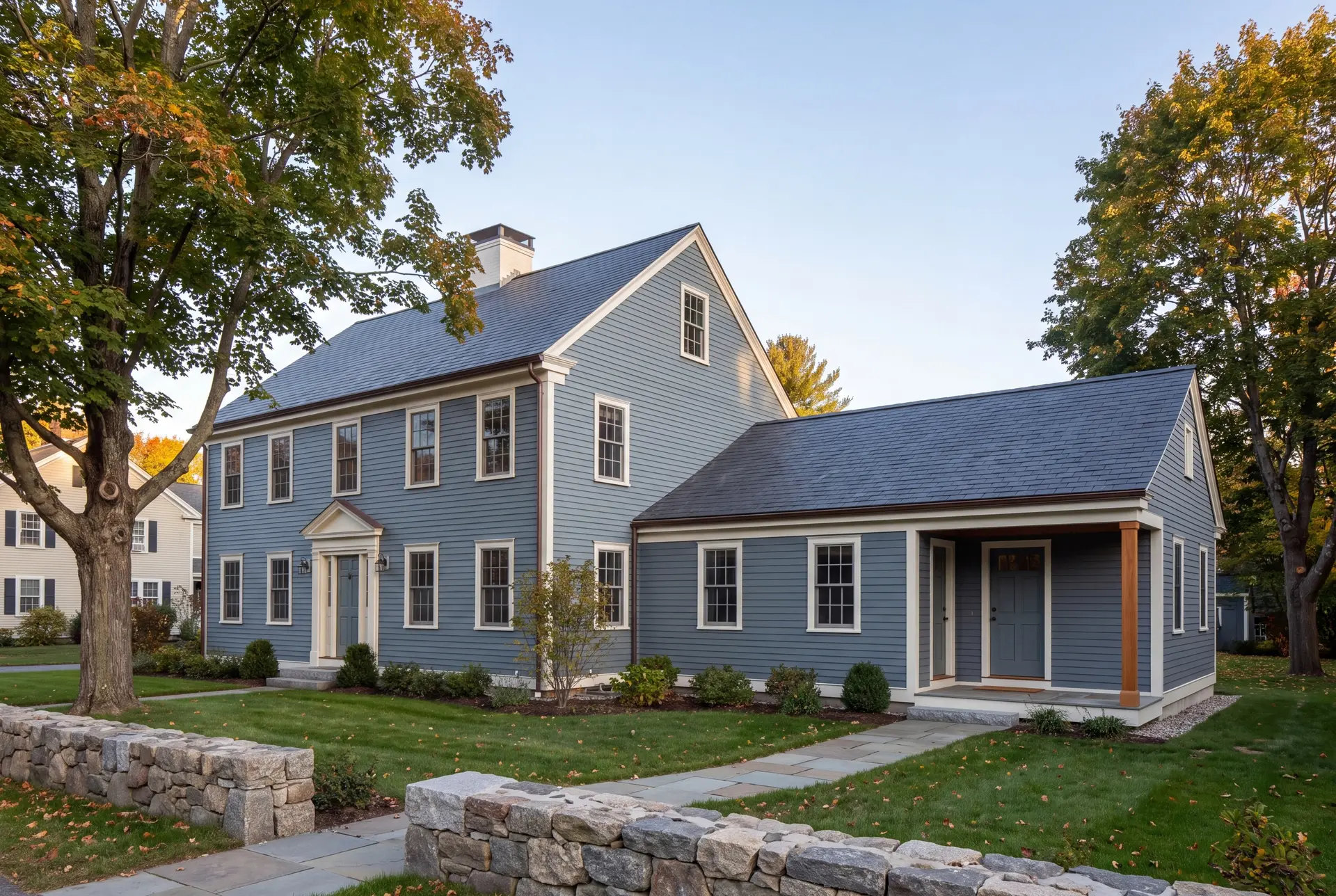 Illustration: An exterior three-quarter view of a blue-gray Colonial house with a new single-story addition, featuring a covered entry and dark gray roofs, set in a landscaped yard with a stone wall