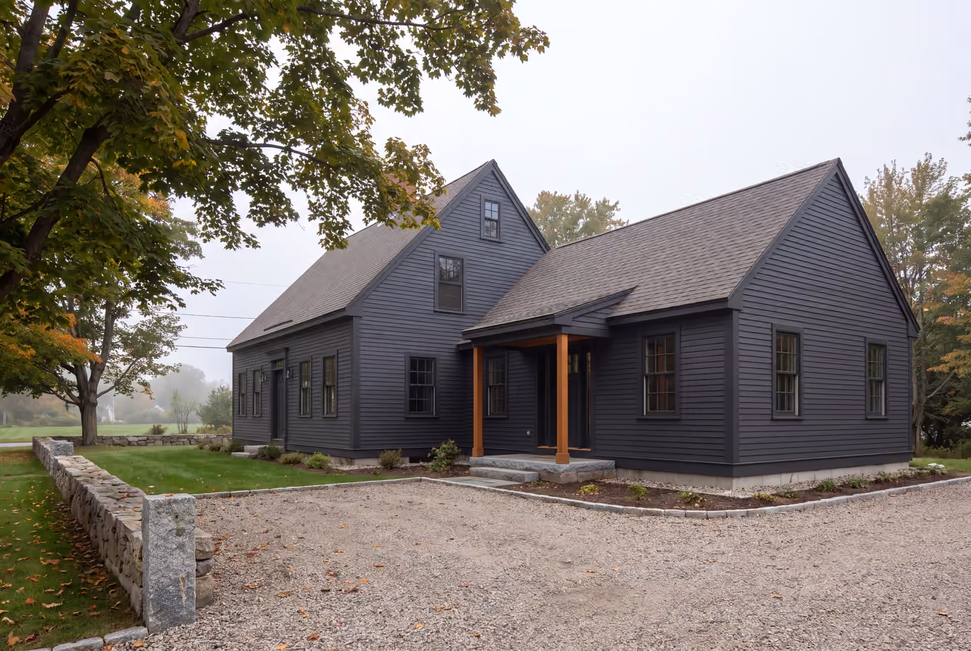 home addition builder in Falmouth, ME: illustrative scene, An exterior view of a dark charcoal clapboard house with a new addition, featuring a covered entry with wooden posts, a gravel driveway, and a stone wall