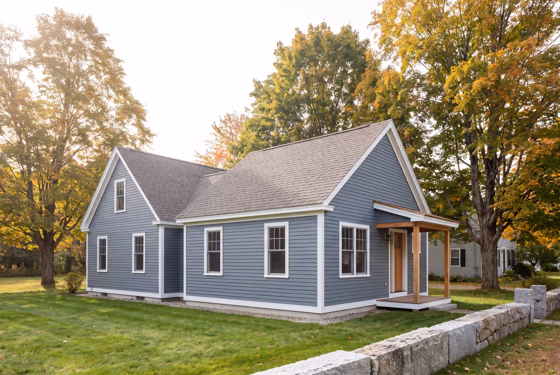 home addition builder in Freeport, ME: illustrative scene, A blue-gray clapboard house with a new single-story addition, featuring a small wooden porch, set in a grassy yard with autumn trees and a stone wall