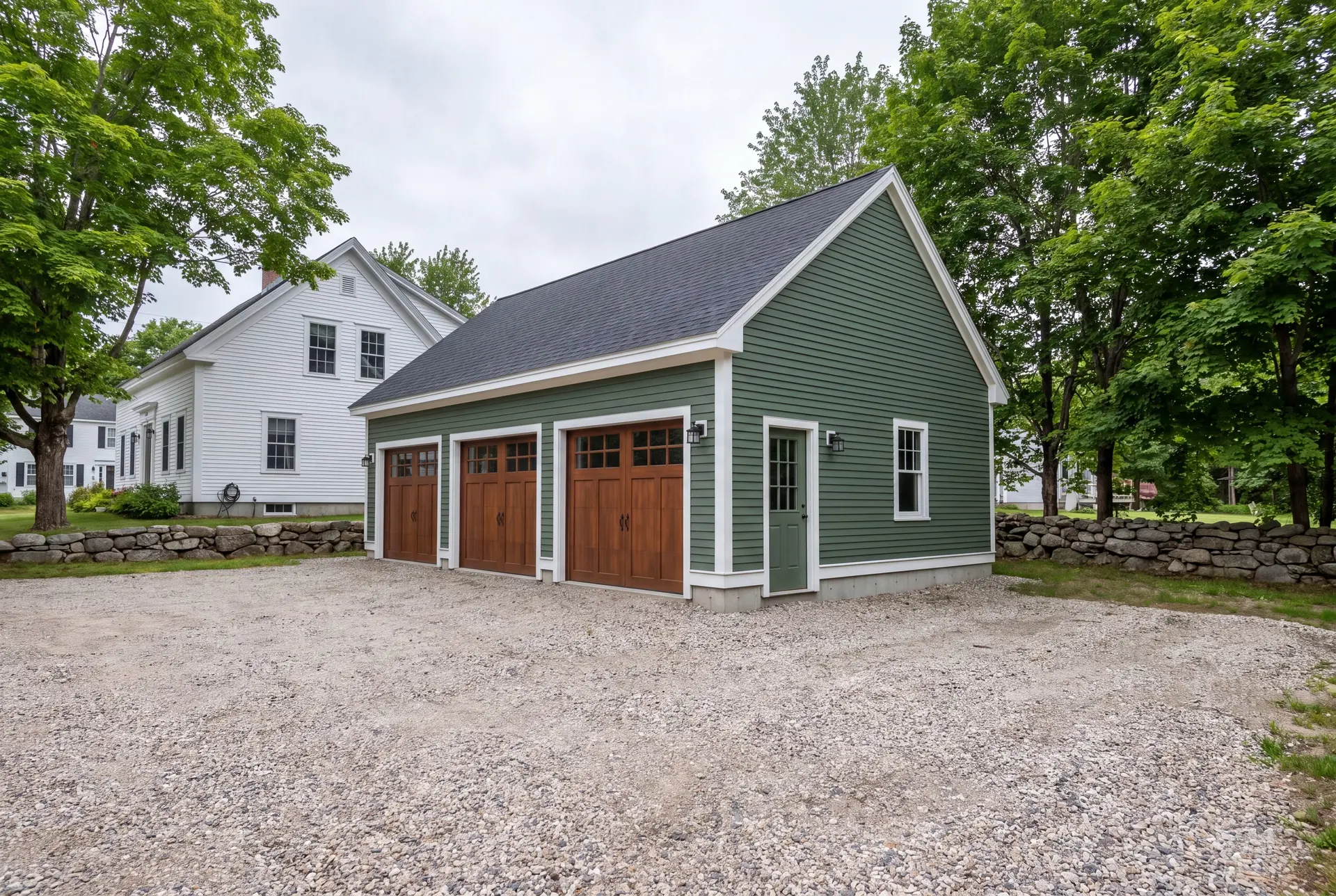 Illustration: A newly built, detached three-bay garage with forest green siding and wooden doors, situated on a gravel driveway next to a white house and surrounded by trees