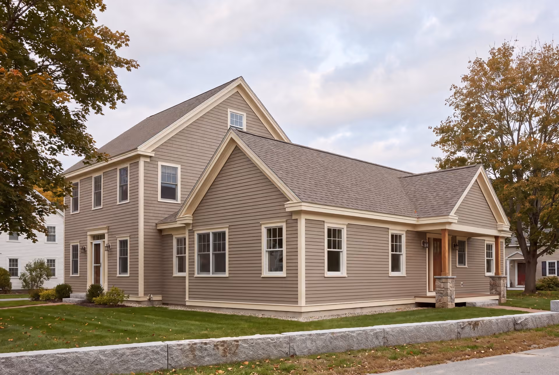 home addition builder in Gorham, ME: illustrative scene, A large, two-story taupe house with a single-story addition, featuring a covered porch with stone and wood posts, surrounded by green lawn and autumn trees under a cloudy sky