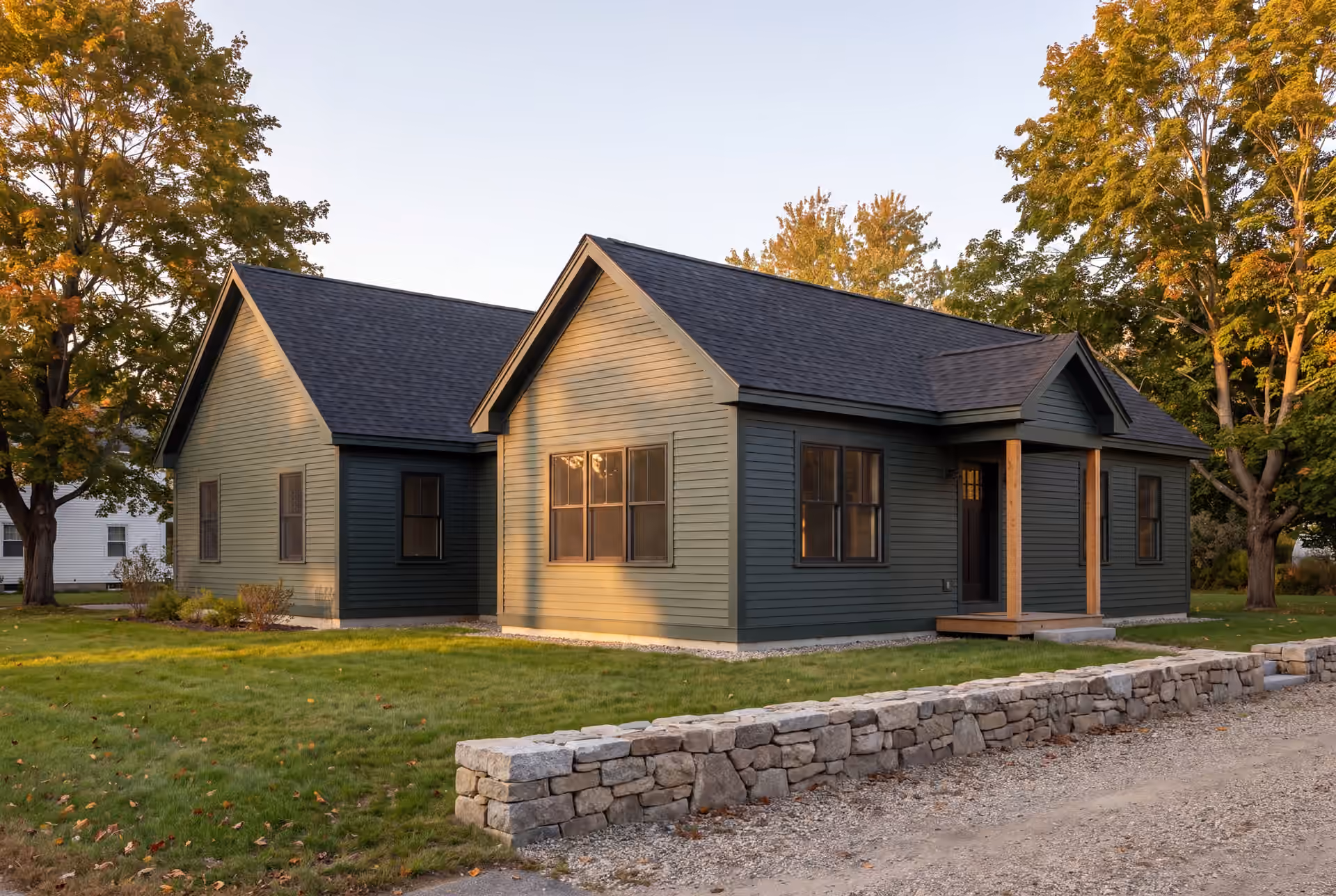 home addition builder in Gray, ME: illustrative scene, A single-story forest green house with a new addition, featuring matching siding and a covered entry with wooden posts, set on a lawn with autumn trees and a stone wall