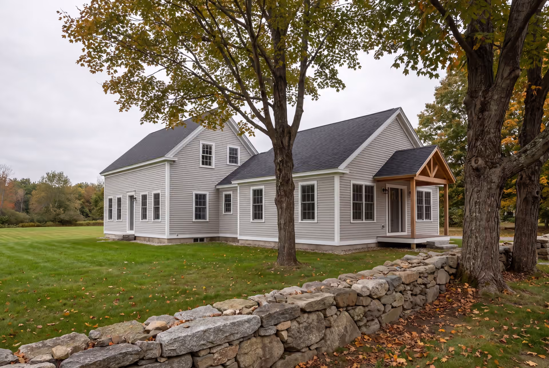 home addition builder in Hollis, ME: illustrative scene, A light gray clapboard farmhouse with a new matching addition, seen from a three-quarter exterior view, surrounded by green grass and trees with autumn leaves, with a stone wall in the foreground