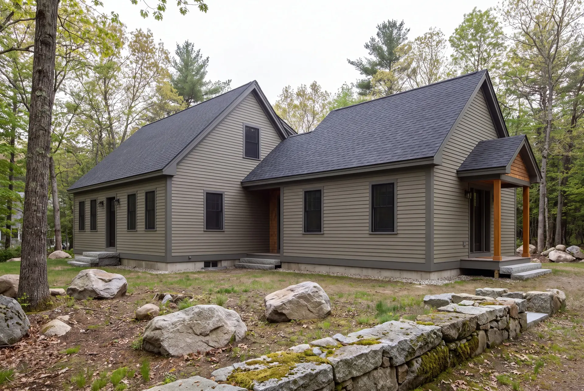 Illustration: A two-part house with dusty olive siding and dark gray roofs, featuring a one-and-a-half-story cottage connected to a single-story addition, surrounded by trees and rocks