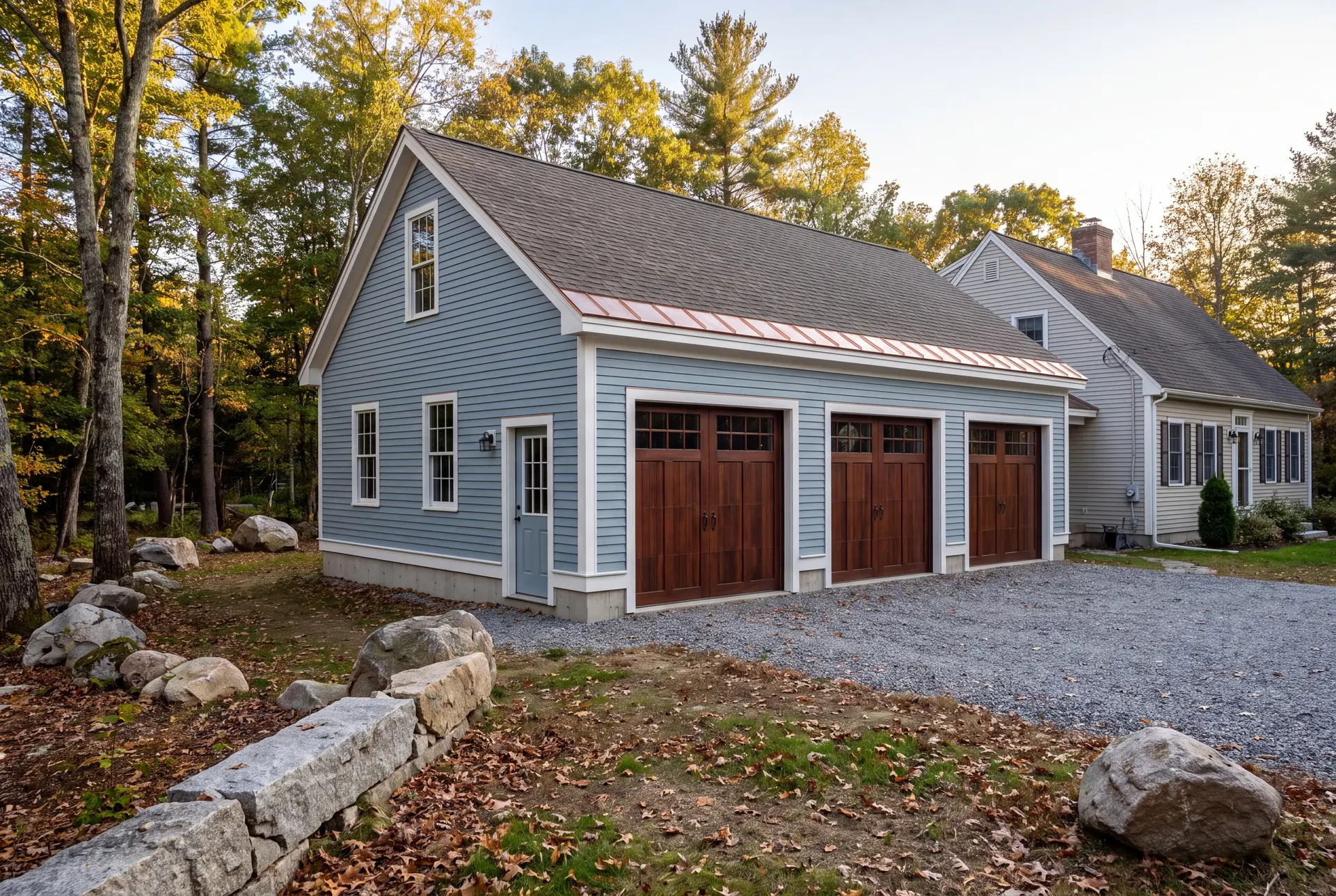 Illustration: A newly built light blue garage with three dark wood doors and a gravel driveway, next to a house surrounded by trees in autumn