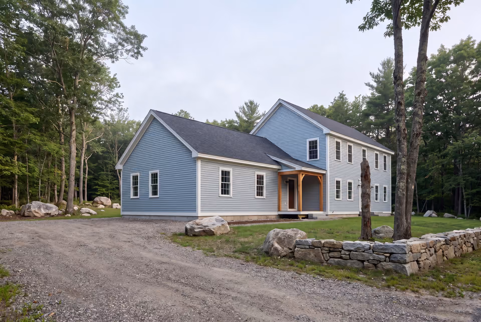 home addition builder in Lyman, ME: illustrative scene, An exterior three-quarter view of a light blue house with a two-story main section and a single-story addition, featuring a covered entry with wood posts, surrounded by a gravel driveway, grass, trees, and boulders