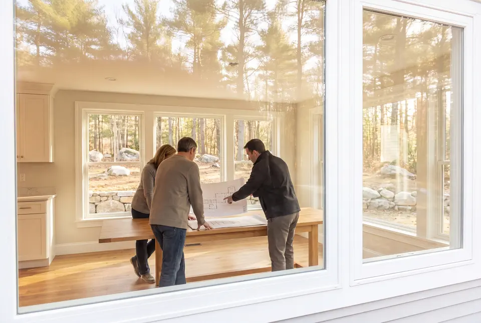 Illustration: Three people, seen from behind, review architectural drawings on a table in a bright, finished living room with large windows overlooking a natural landscape