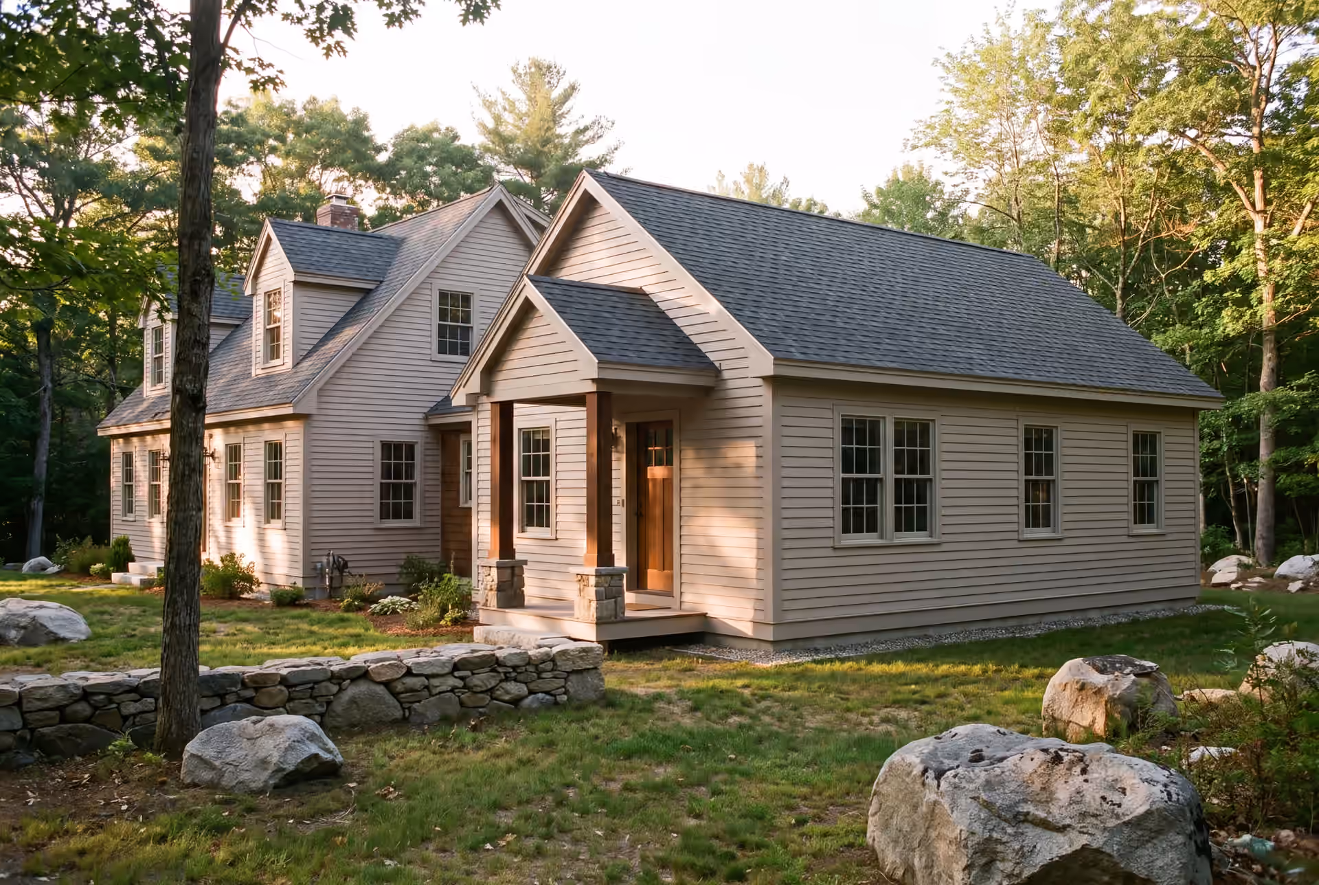 home addition builder in Sanford, ME: illustrative scene, An exterior view of a light-colored house with a new addition, surrounded by a green lawn, trees, and large rocks