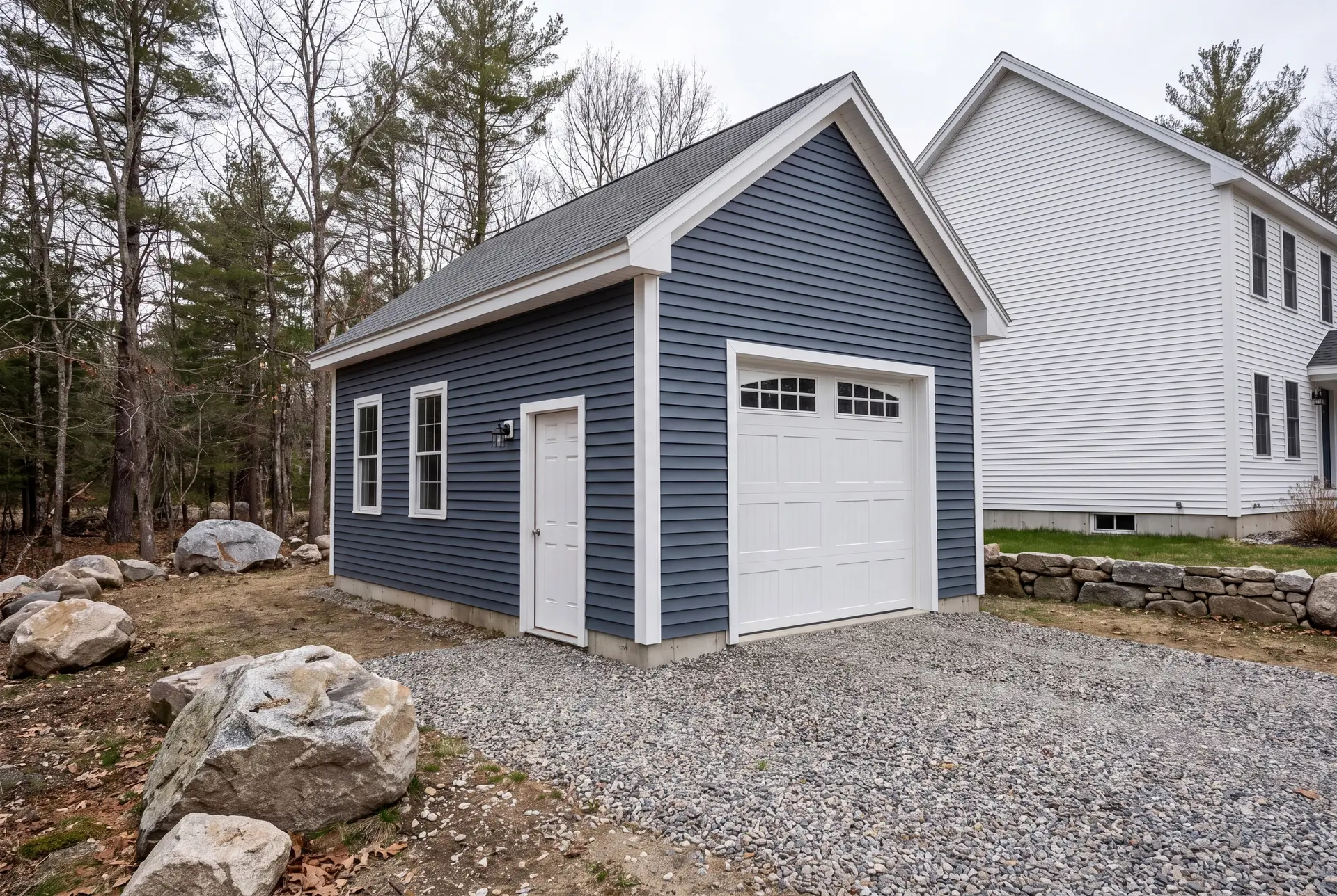 Illustration: A newly built, single-bay garage with deep slate blue siding and white trim, a white carriage-style garage door, and a gravel driveway