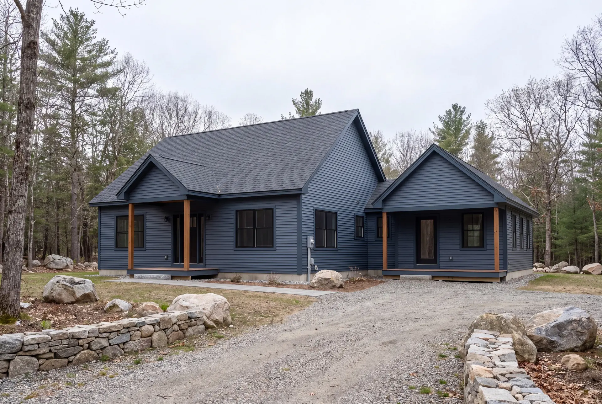 Illustration: An exterior three-quarter view of a dark blue single-story house with a new addition, featuring gabled roofs, dark windows, and wooden support posts for the covered entries, set against a backdrop of trees and a gravel driveway