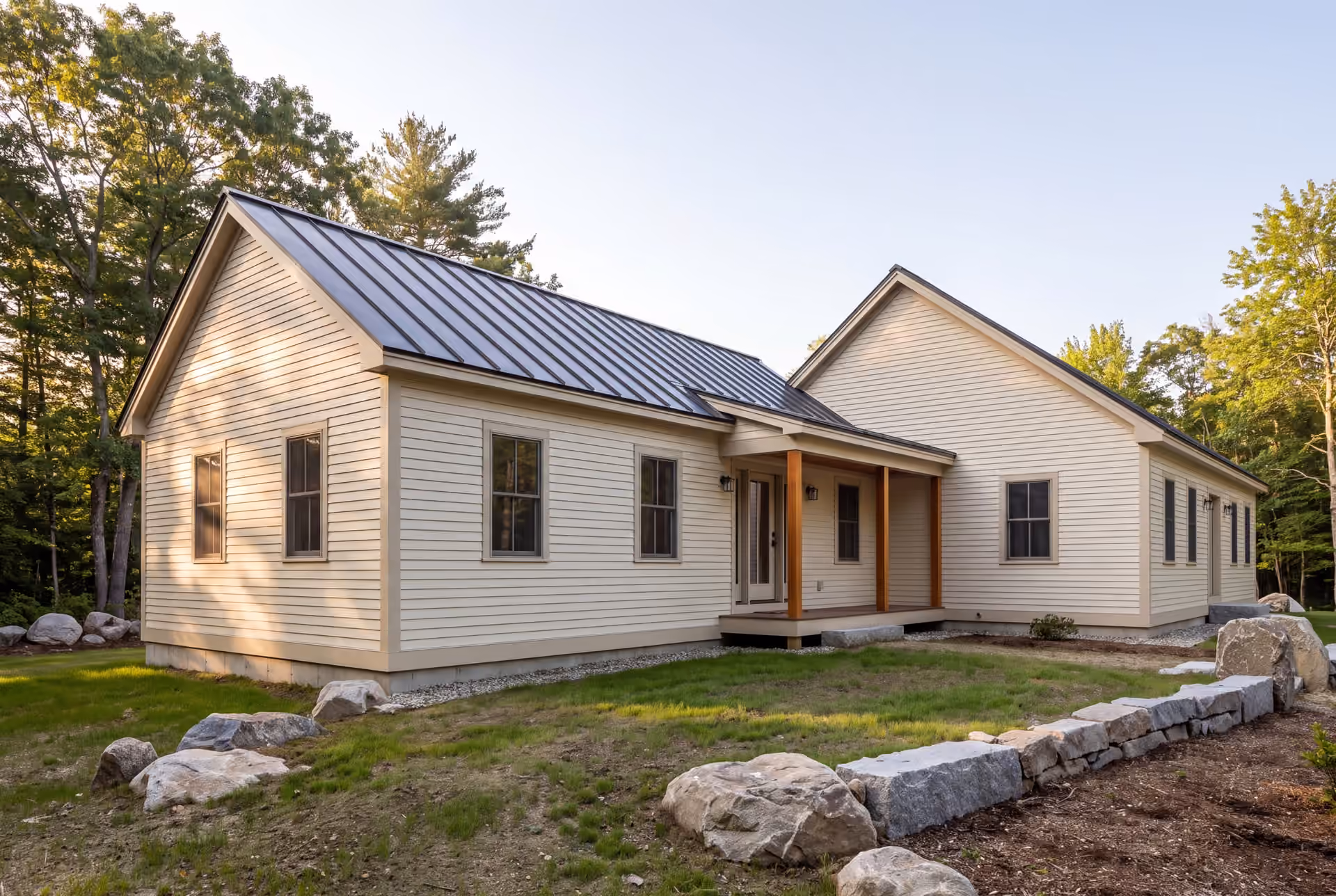 home addition builder in South Portland, ME: illustrative scene, An exterior view of a single-story house with a new addition, featuring light-colored siding and a dark metal roof, surrounded by grass and trees