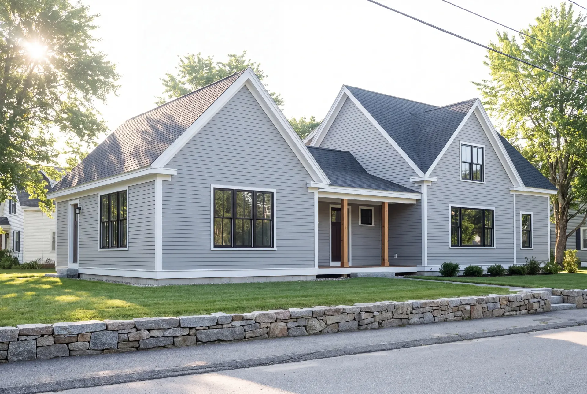 Illustration: An exterior three-quarter view of a gray clapboard house with a new addition, featuring gabled roofs, black-framed windows, and a covered entry with wooden posts, set on a green lawn behind a stone wall