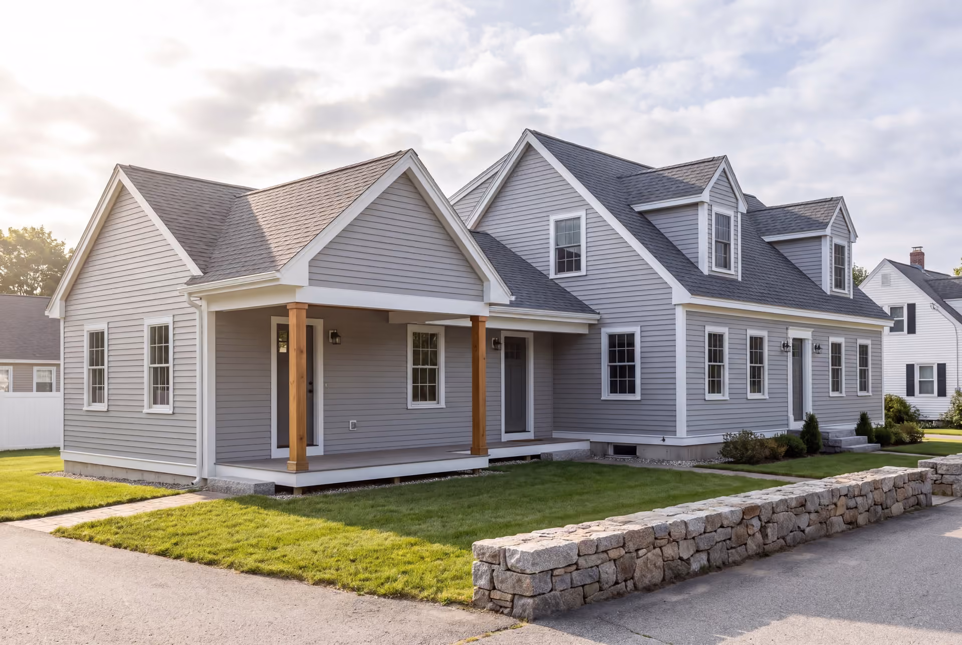 home addition builder in Westbrook, ME: illustrative scene, A light gray clapboard house with a new single-story addition, featuring a covered porch with wooden posts, set against a partly cloudy sky