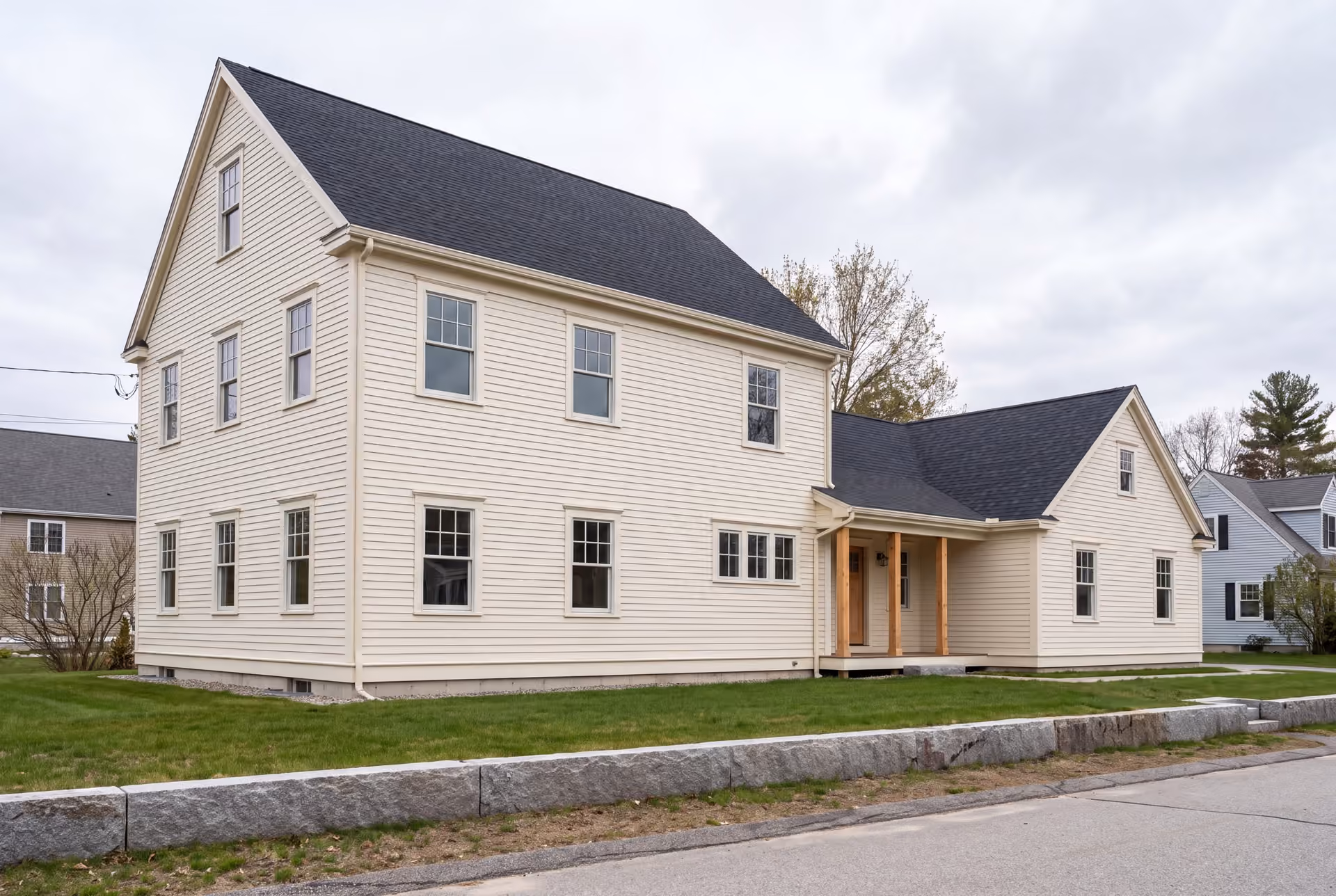 home addition builder in Windham, ME: illustrative scene, A cream-colored two-story Colonial house with a new single-story addition, featuring a covered entry with timber posts, on a green lawn under an overcast sky