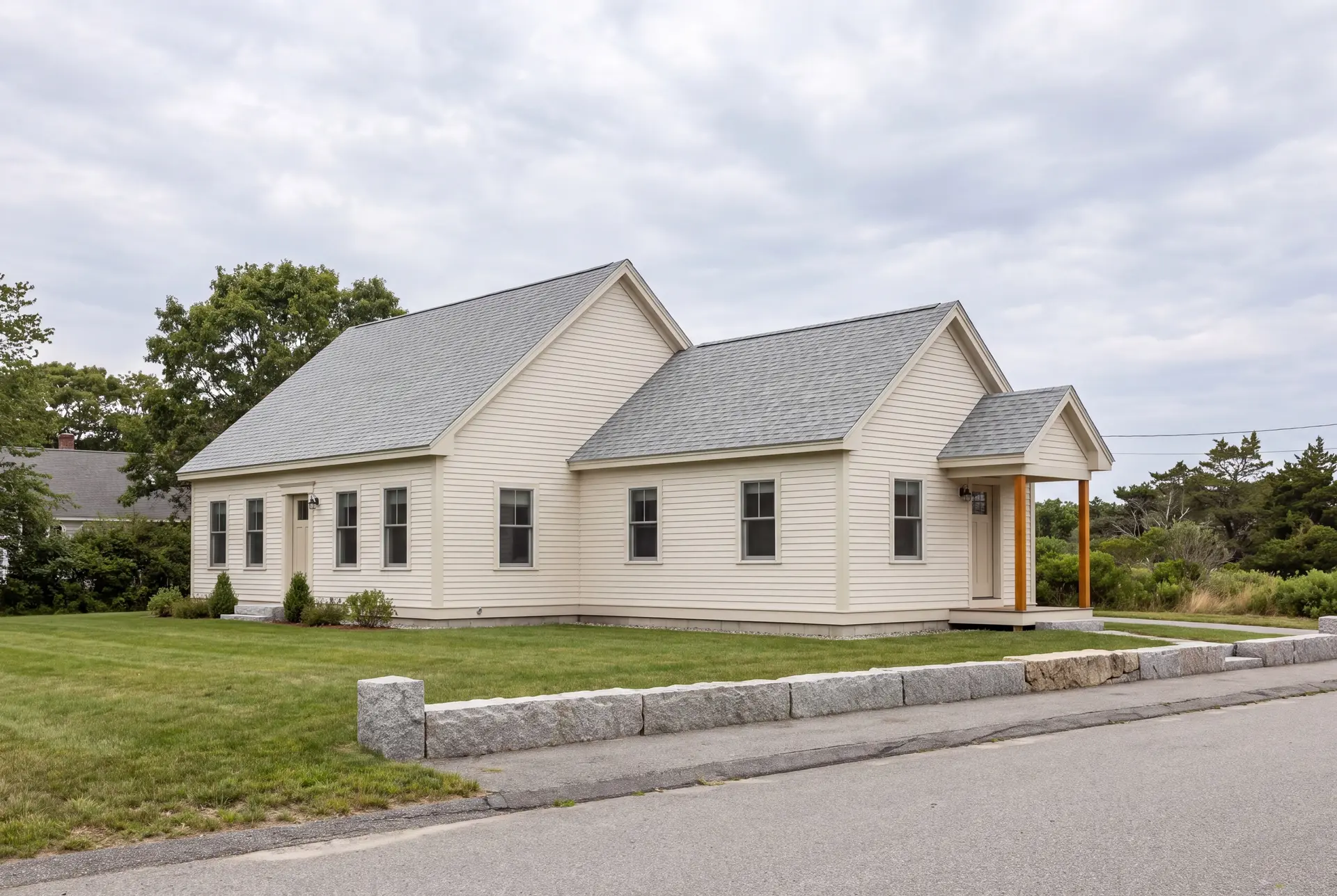 Illustration: A cream-colored single-story house with a new addition, featuring a gray shingle roof and a small covered entry with wooden posts, set on a green lawn next to a stone wall and road