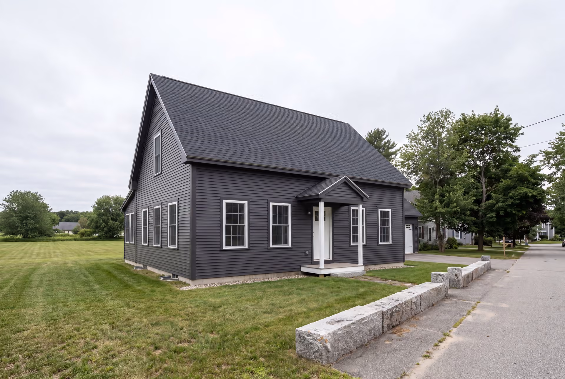 custom home builder in Biddeford, ME: illustrative scene, A dark charcoal cottage with a gabled roof and white-trimmed windows, viewed from the front and side, next to a road and lawn