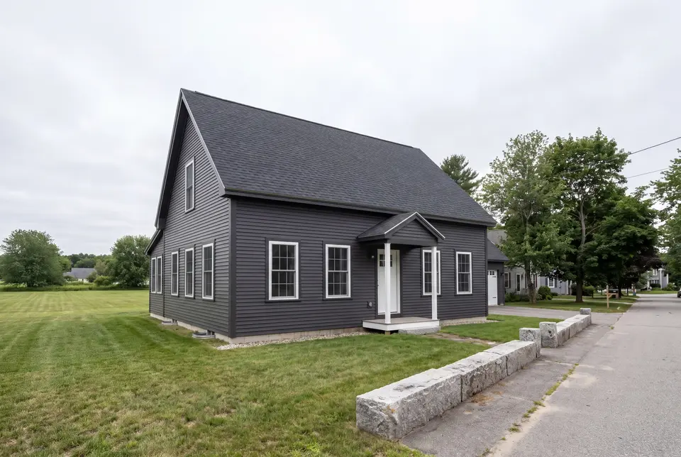 custom home builder in Biddeford, ME: illustrative scene, A dark charcoal cottage with a gabled roof and white-trimmed windows, viewed from the front and side, next to a road and lawn