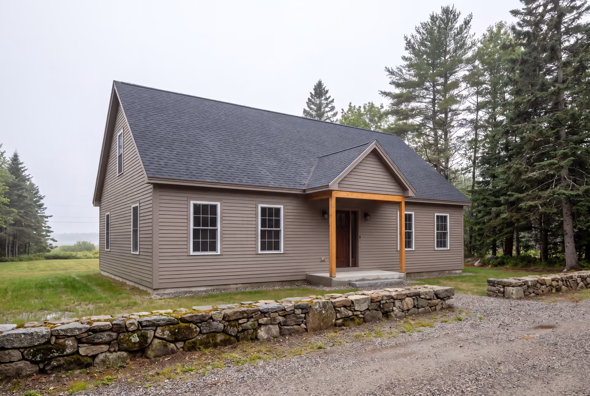 custom home builder in Cape Elizabeth, ME: illustrative scene, A newly built single-story house with taupe siding, a dark roof, and a wood-post covered entry, viewed from the front and side, with a stone wall and gravel driveway in the foreground