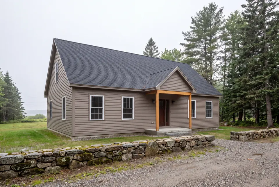 custom home builder in Cape Elizabeth, ME: illustrative scene, A newly built single-story house with taupe siding, a dark roof, and a wood-post covered entry, viewed from the front and side, with a stone wall and gravel driveway in the foreground
