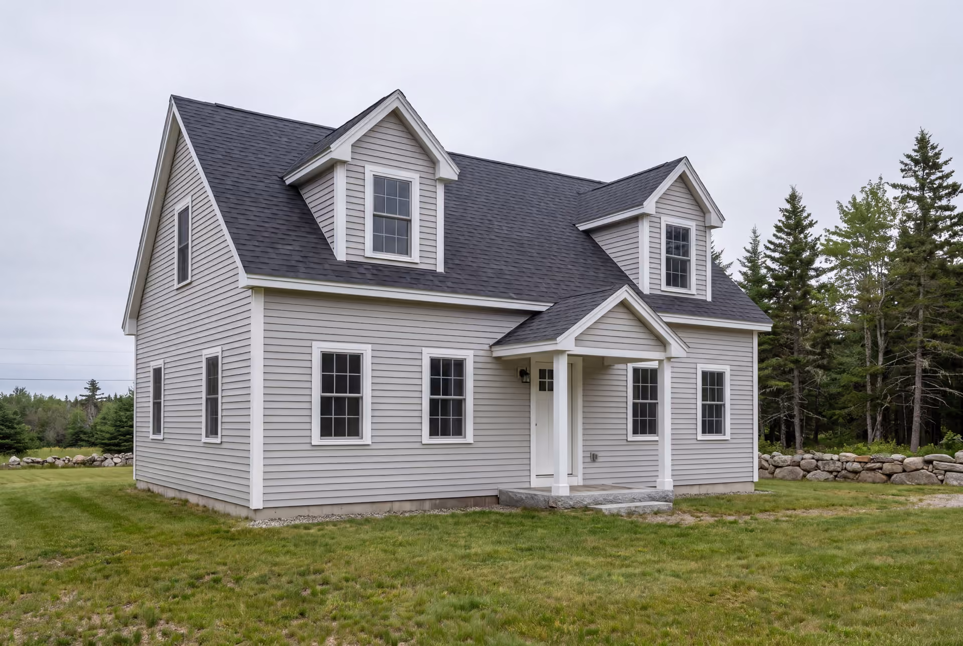 custom home builder in Dayton, ME: illustrative scene, A newly built Cape house with light gray siding, a dark roof, and two dormers, set on a green lawn with trees and a stone wall in the background