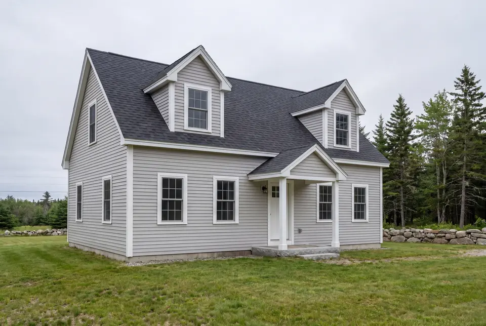 custom home builder in Dayton, ME: illustrative scene, A newly built Cape house with light gray siding, a dark roof, and two dormers, set on a green lawn with trees and a stone wall in the background
