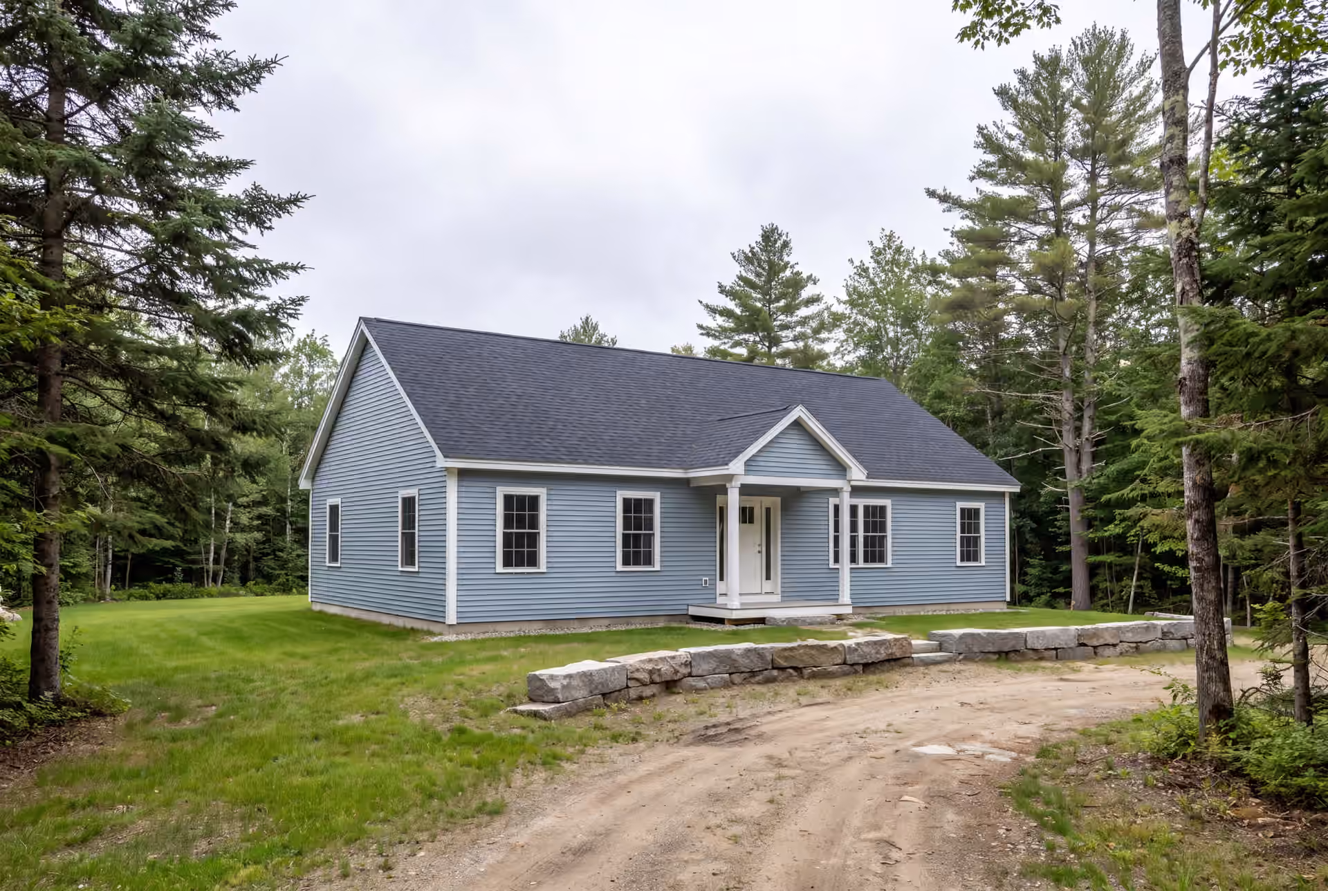custom home builder in Freeport, ME: illustrative scene, A light blue ranch house with a charcoal roof and white trim, surrounded by trees and grass, with a dirt driveway and stone wall in the foreground