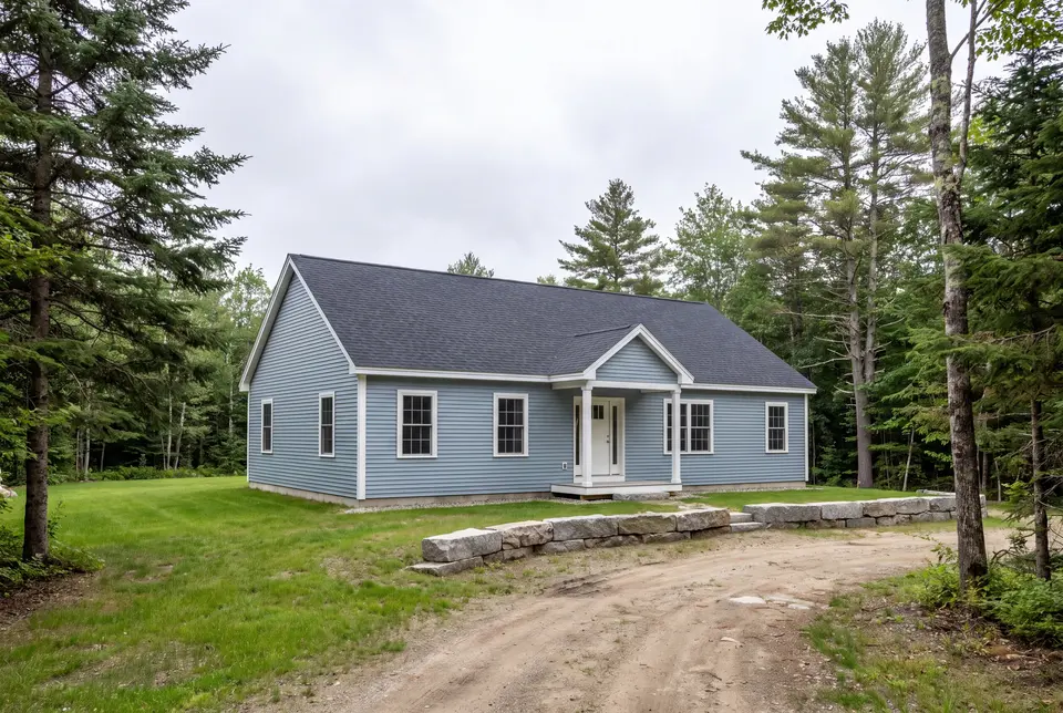 custom home builder in Freeport, ME: illustrative scene, A light blue ranch house with a charcoal roof and white trim, surrounded by trees and grass, with a dirt driveway and stone wall in the foreground