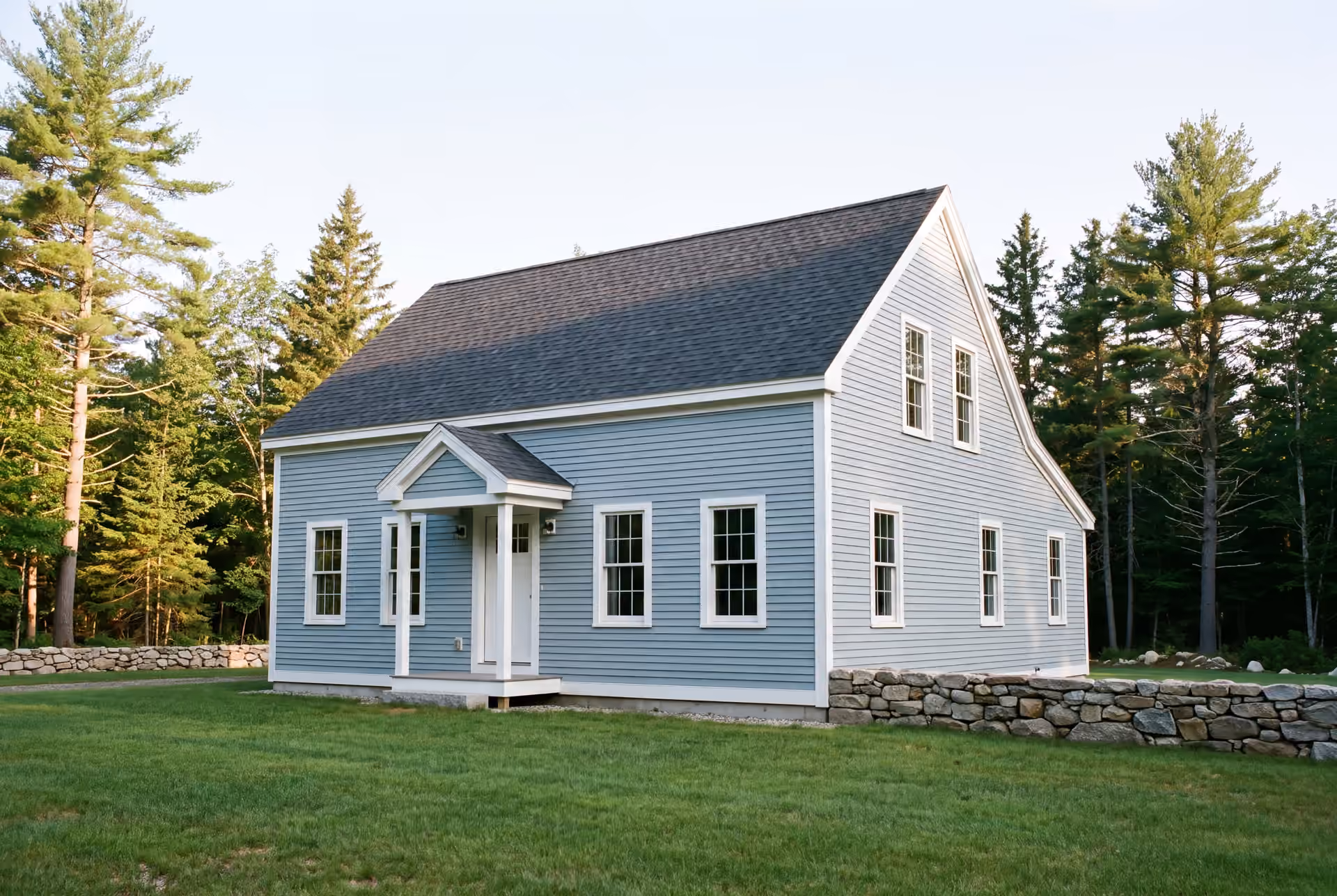 custom home builder in Gorham, ME: illustrative scene, A light blue New England saltbox house with a dark roof and white trim, surrounded by green grass and pine trees, with a stone wall in the foreground
