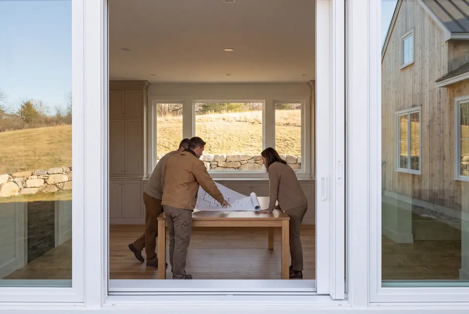 Illustration: Three people, a builder and two homeowners, reviewing architectural drawings on a table in a modern living room with large windows overlooking a rural landscape
