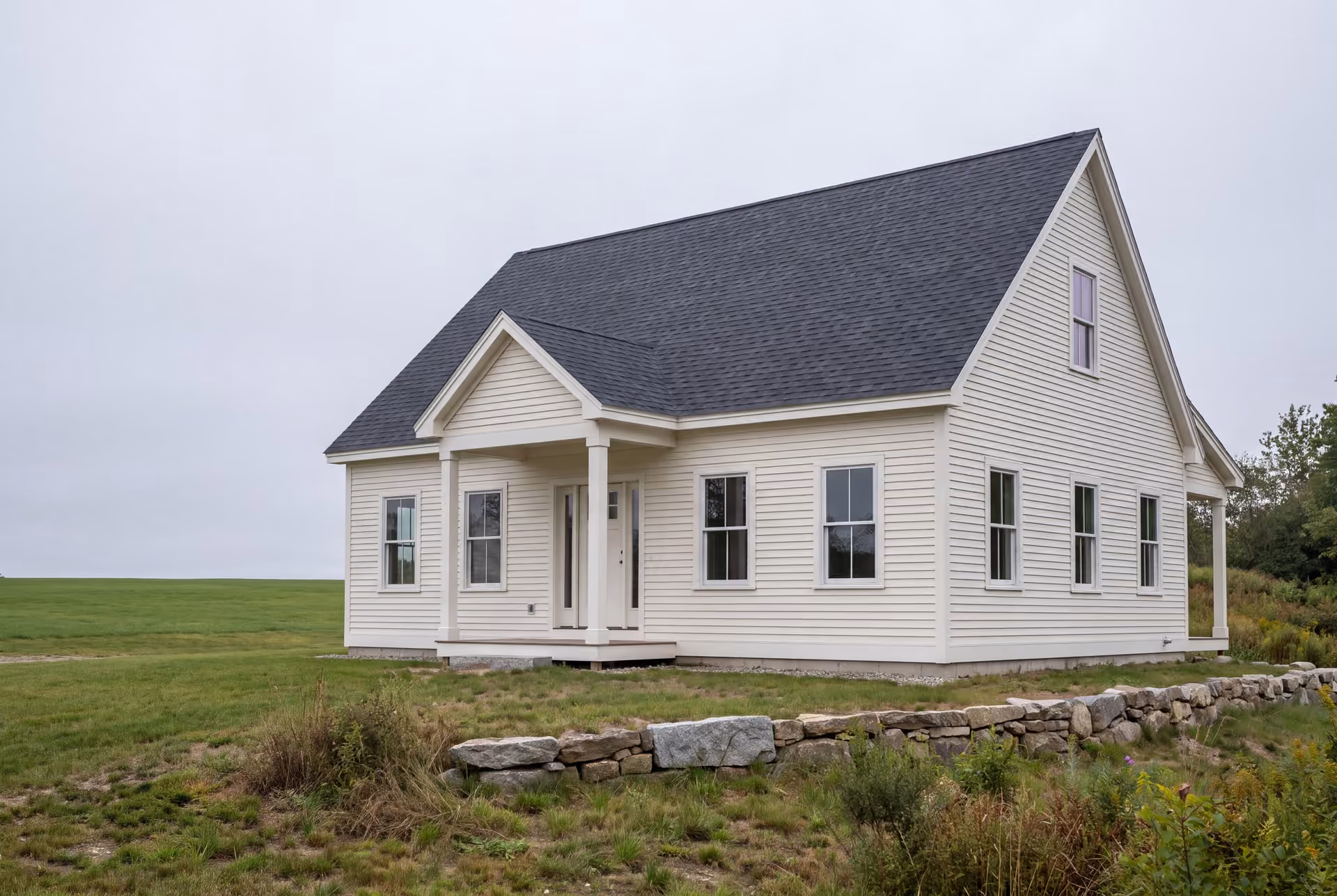 custom home builder in Gray, ME: illustrative scene, A newly built white clapboard cottage with a dark gray roof, white-trimmed windows, and a covered porch, situated in a green field under a gray sky
