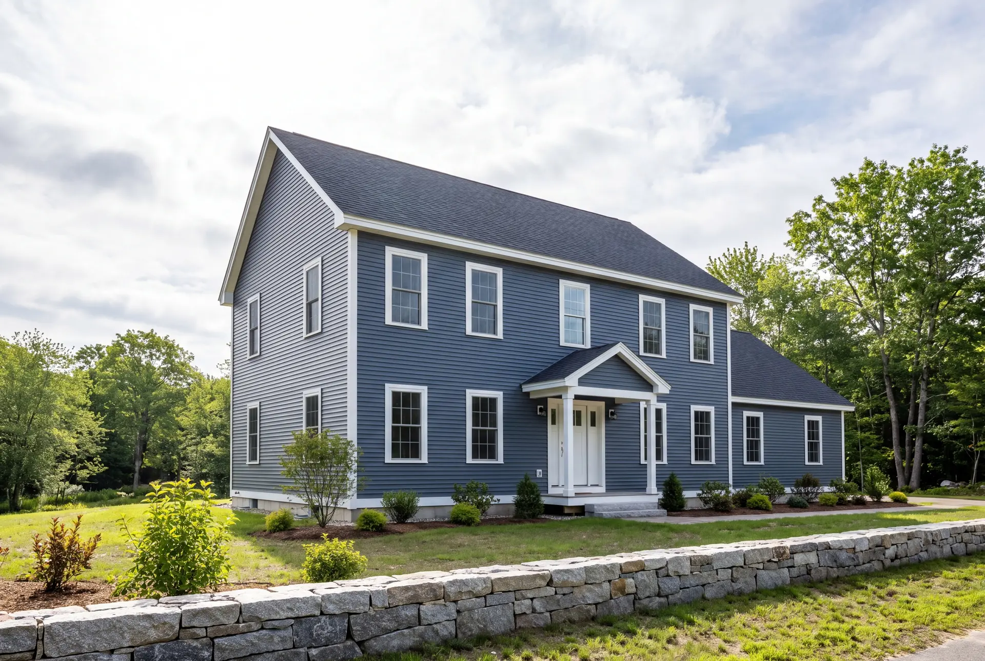 Illustration: A newly built two-story Colonial house with blue siding, a dark roof, and white trim, viewed from the front and side, with a stone wall in the foreground
