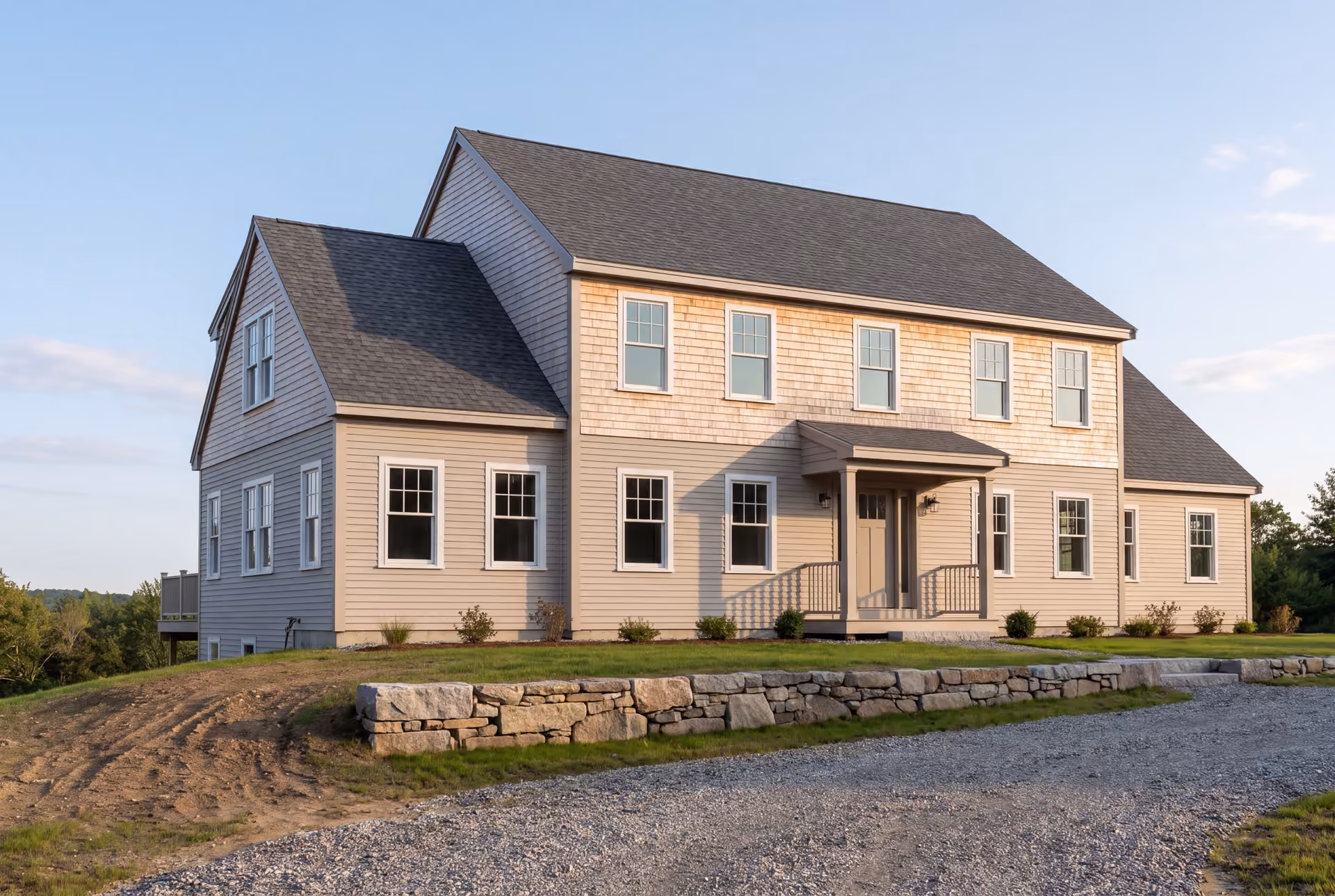 custom home builder in Hollis, ME: illustrative scene, A newly built two-story shingle-style house with light-colored siding, a dark gray roof, and white-trimmed windows, seen from a three-quarter view with a gravel driveway and stone wall in the foreground