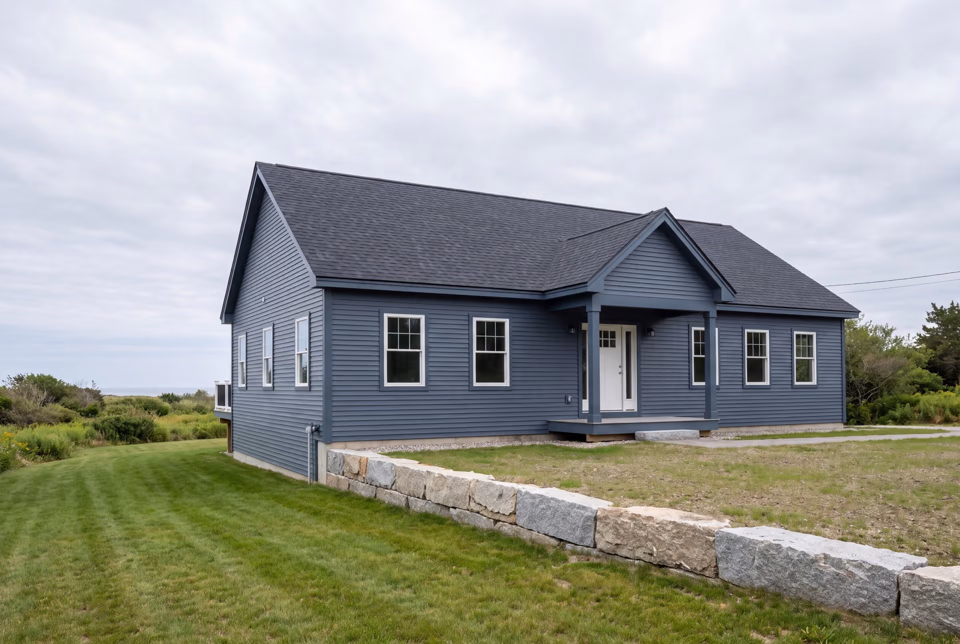 custom home builder in Kennebunk, ME: illustrative scene, A newly built single-story house with blue siding, a dark roof, and a covered porch, set in a grassy landscape with a stone wall
