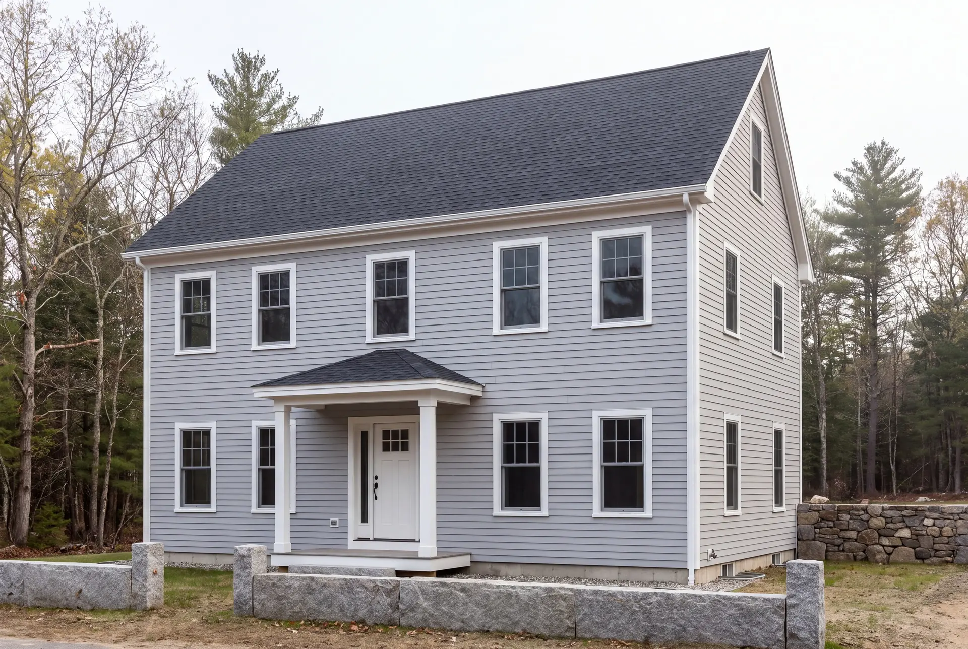 Illustration: A newly built two-story gray house with a dark roof and white trim, seen from a three-quarter angle, surrounded by trees and a stone wall