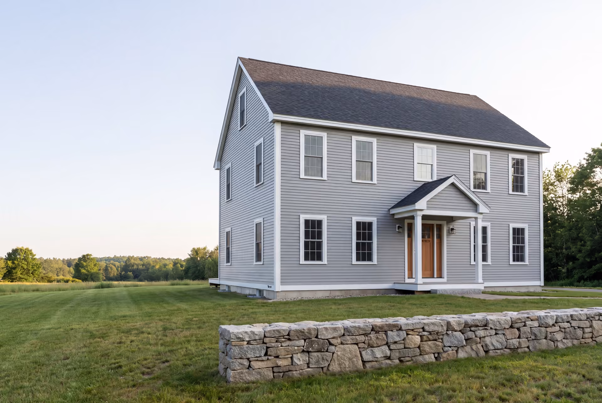 custom home builder in Saco, ME: illustrative scene, A newly built two-story gray Colonial house with a dark roof and white trim, seen from a three-quarter angle, with a stone wall in the foreground and a grassy field extending to a tree line
