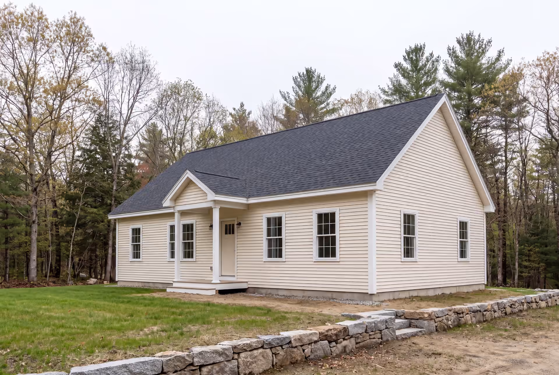 custom home builder in Sanford, ME: illustrative scene, A newly built cream-colored ranch house with a dark gray roof and white trim, seen from a three-quarter angle, with a stone wall in the foreground and trees in the background