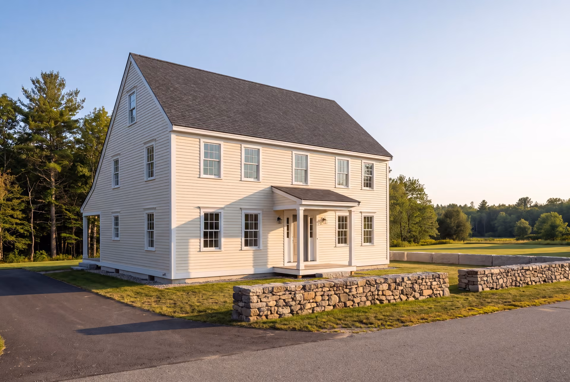custom home builder in Scarborough, ME: illustrative scene, A newly built, traditional New England saltbox house with cream siding and a dark roof, viewed from an angle showing the front and side, with a stone wall in the foreground