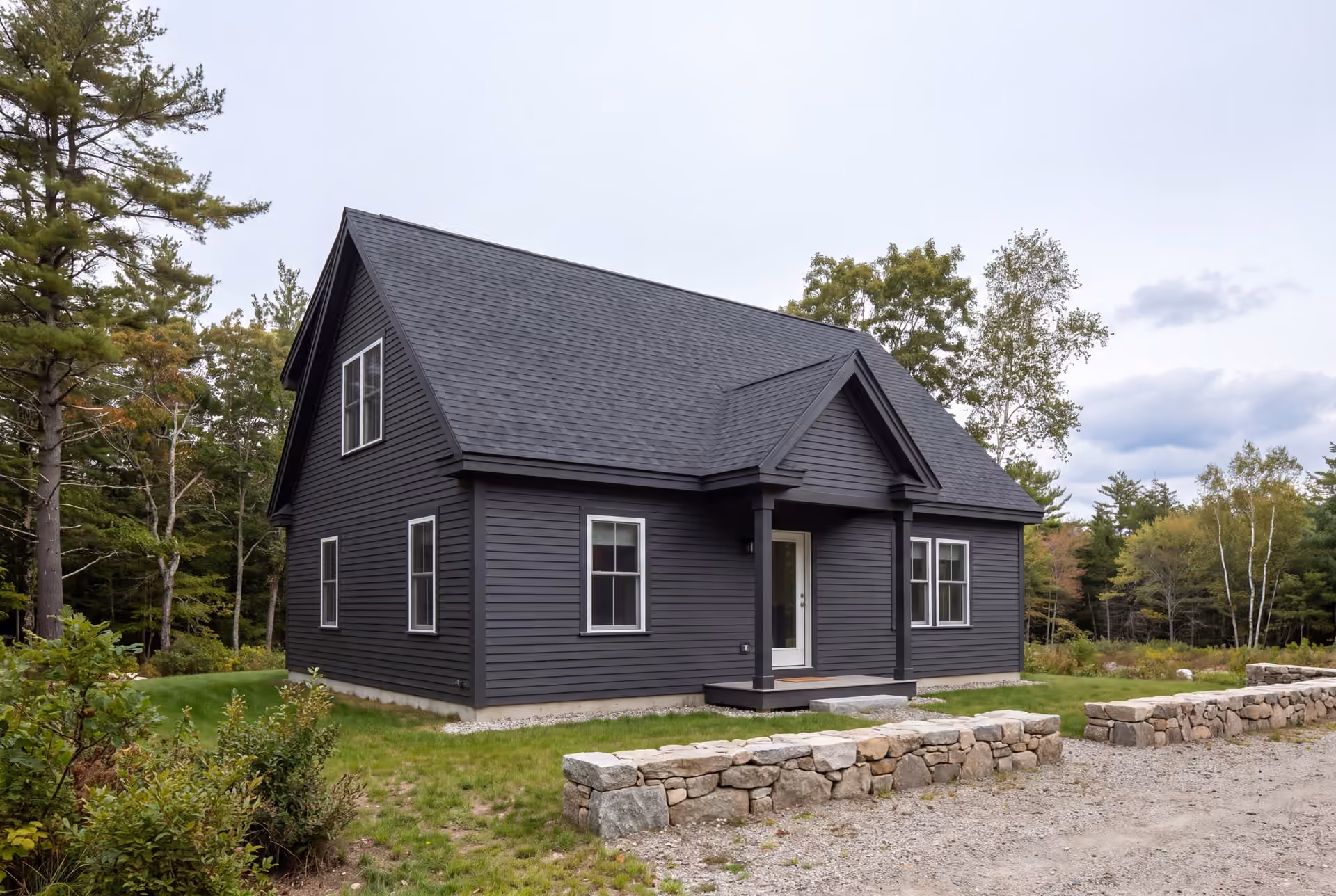 custom home builder in South Portland, ME: illustrative scene, A dark charcoal cottage with a gabled roof and white-trimmed windows, set in a natural landscape with trees and a gravel driveway